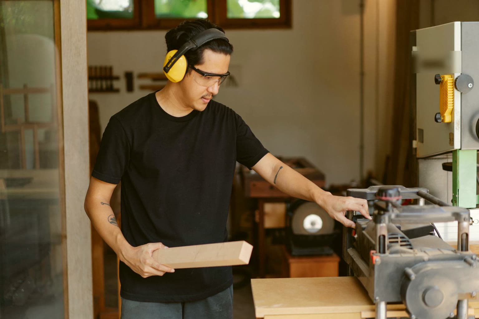 Woodworker feeding a board into a planer in a sunlit workshop