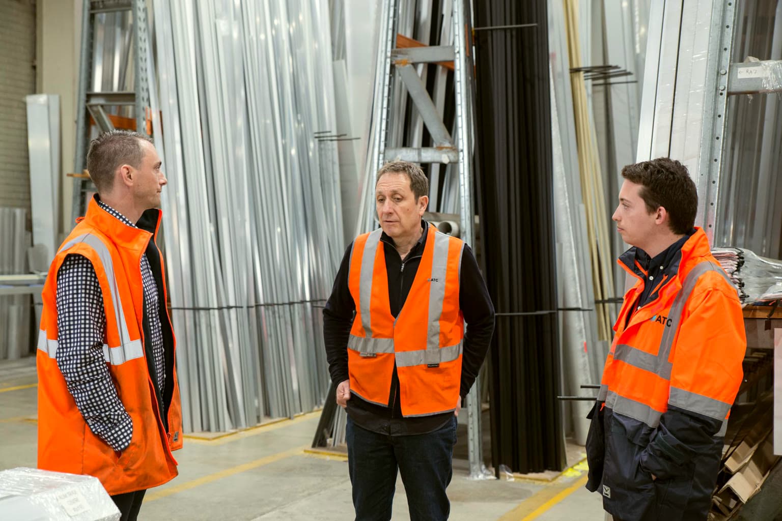 Three warehouse employees in orange safety vests talking on the shop floor